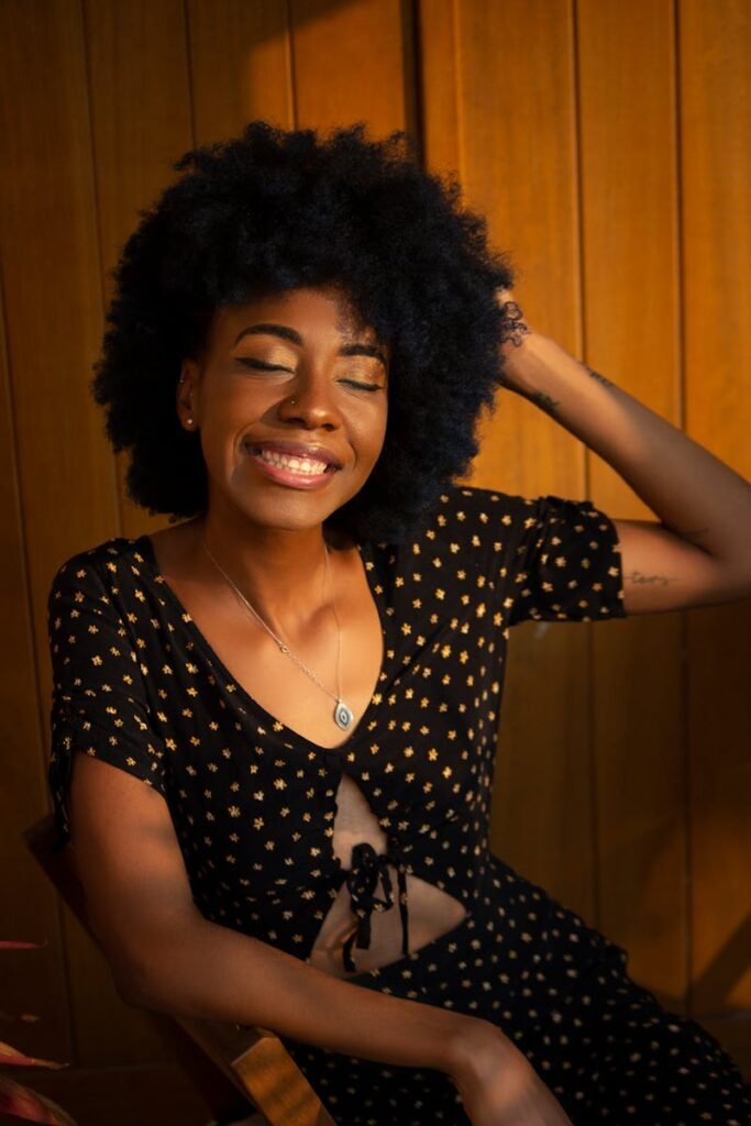 A beautiful African woman with an afro hairstyle smiling indoors, wearing a stylish dress and necklace.